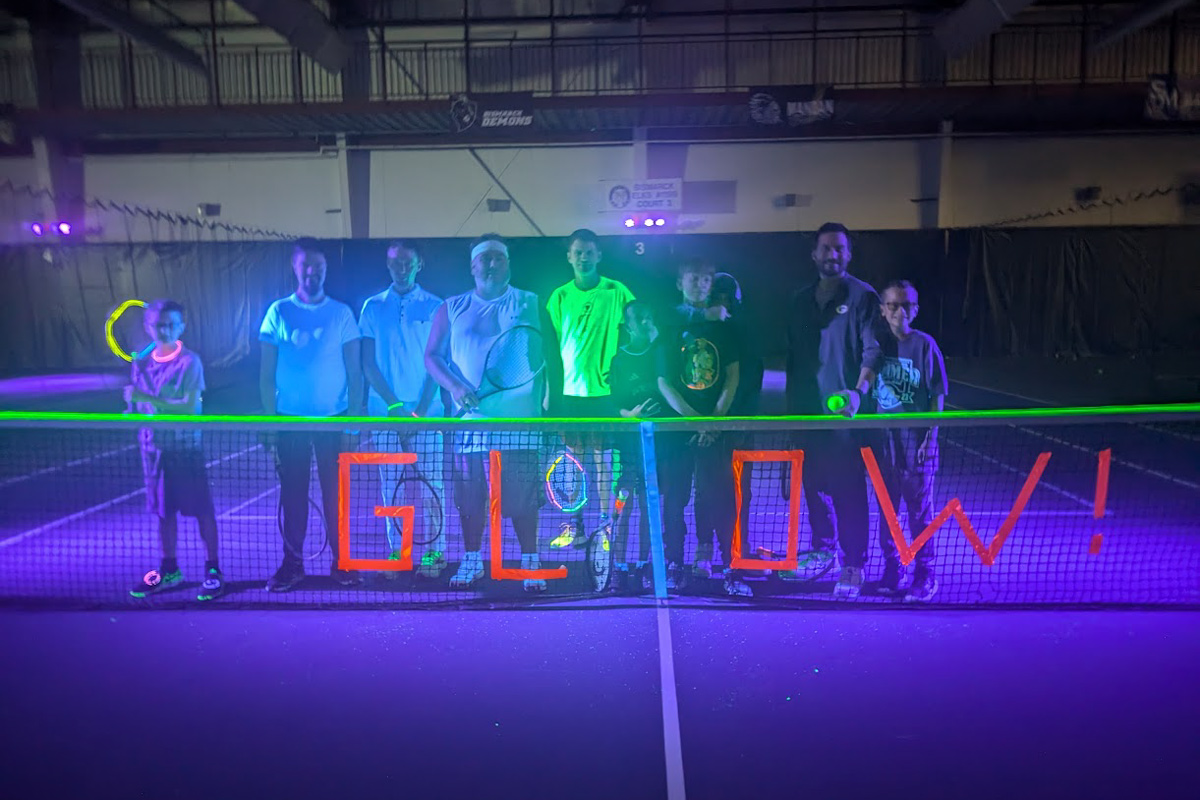 group photo of blacklight tennis participants in front of a tennis net that says "Glow!" in glow in the dark duct tape